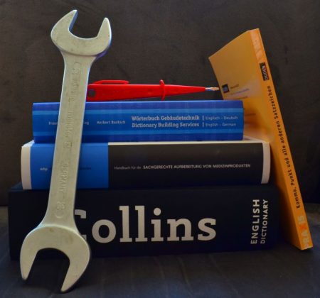Ein Stapel Fach- und Wörterbücher, dazu Werkzeug. Diese Kombination steht symbolisch für Sprache und Technik.