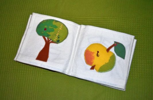 Ein Stoffbuch für Kleinkinder, die aufgeschlagenen Seiten zeigen einen Baum und einen Apfel