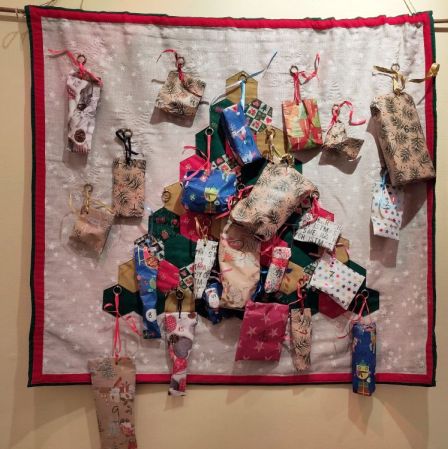 An einem weihnachtlichen Wandbehang hängen viele kleine Adventskalenderpäckchen.